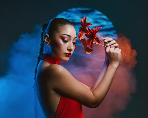 A woman in red dress holding a flower in front of a smokey background