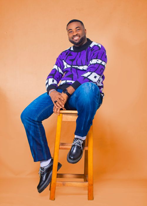 Free Smiling man in a colorful sweater and jeans sitting on a stool with a vibrant yellow backdrop. Stock Photo