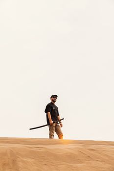 A lone man with a stick walks on a desert dune under a clear sky, symbolizing adventure and solitude.