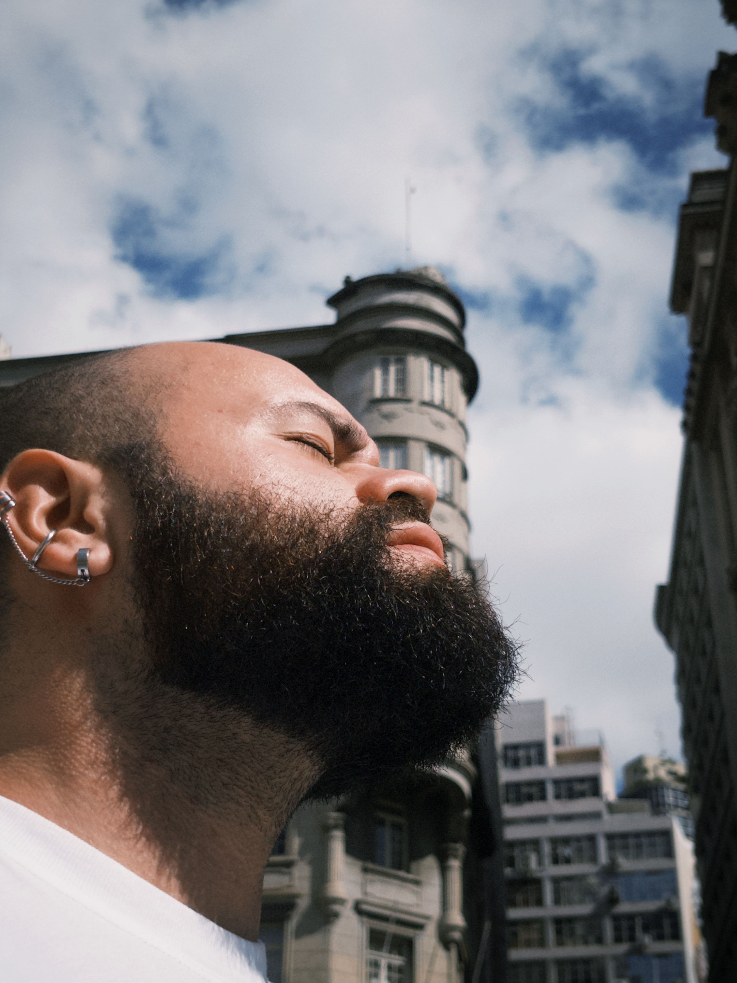 Close-up portrait of a man with a beard and earrings, eyes closed, enjoying the sun against a city backdrop.