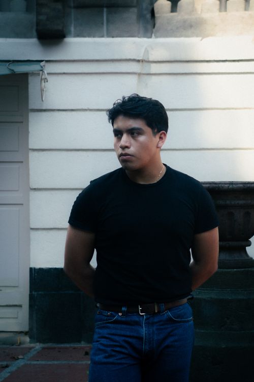 Free A young man in a black t-shirt stands against an urban backdrop, exuding a fashionable and thoughtful look. Stock Photo