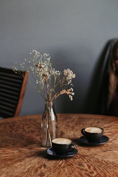 木桌上放着两个泡沫艺术卡布奇诺杯和一个精致的花瓶