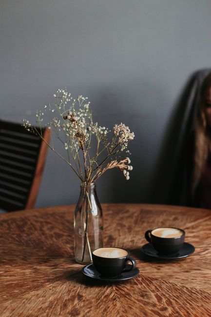 Two cappuccino cups with frothy art on a wooden table, accompanied by a delicate flower vase.