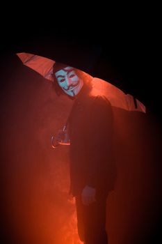 A mysterious figure wearing a Guy Fawkes mask stands under an umbrella in moody lighting.
