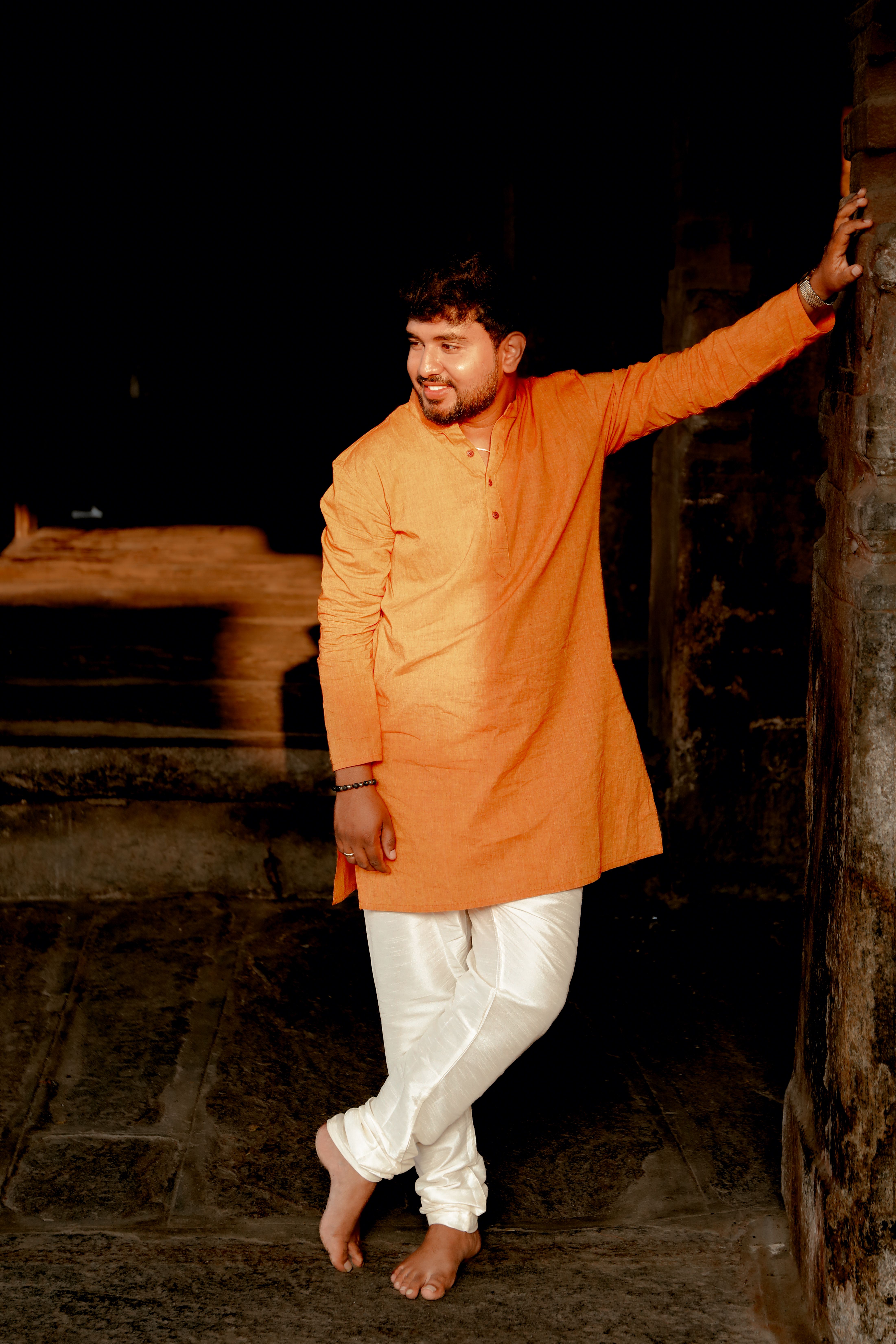 A man wearing traditional Indian clothing, standing in a dimly lit setting, with an orange kurta and white pajama.