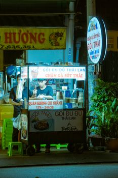 A street food vendor serves Thai cuisine from an illuminated cart on a bustling urban street at night.