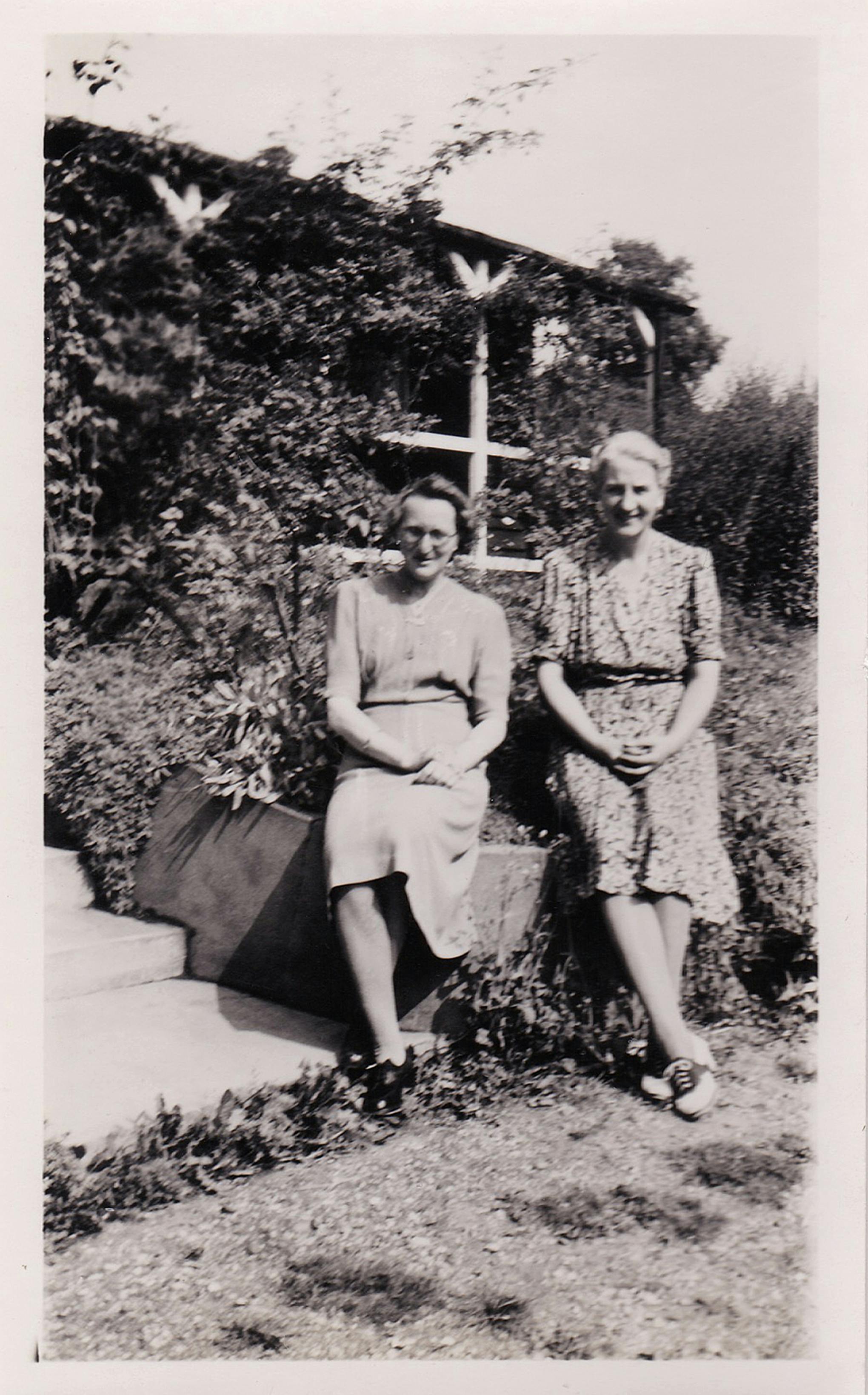 A black and white photo of two women sitting outdoors in a garden, showcasing vintage fashion.