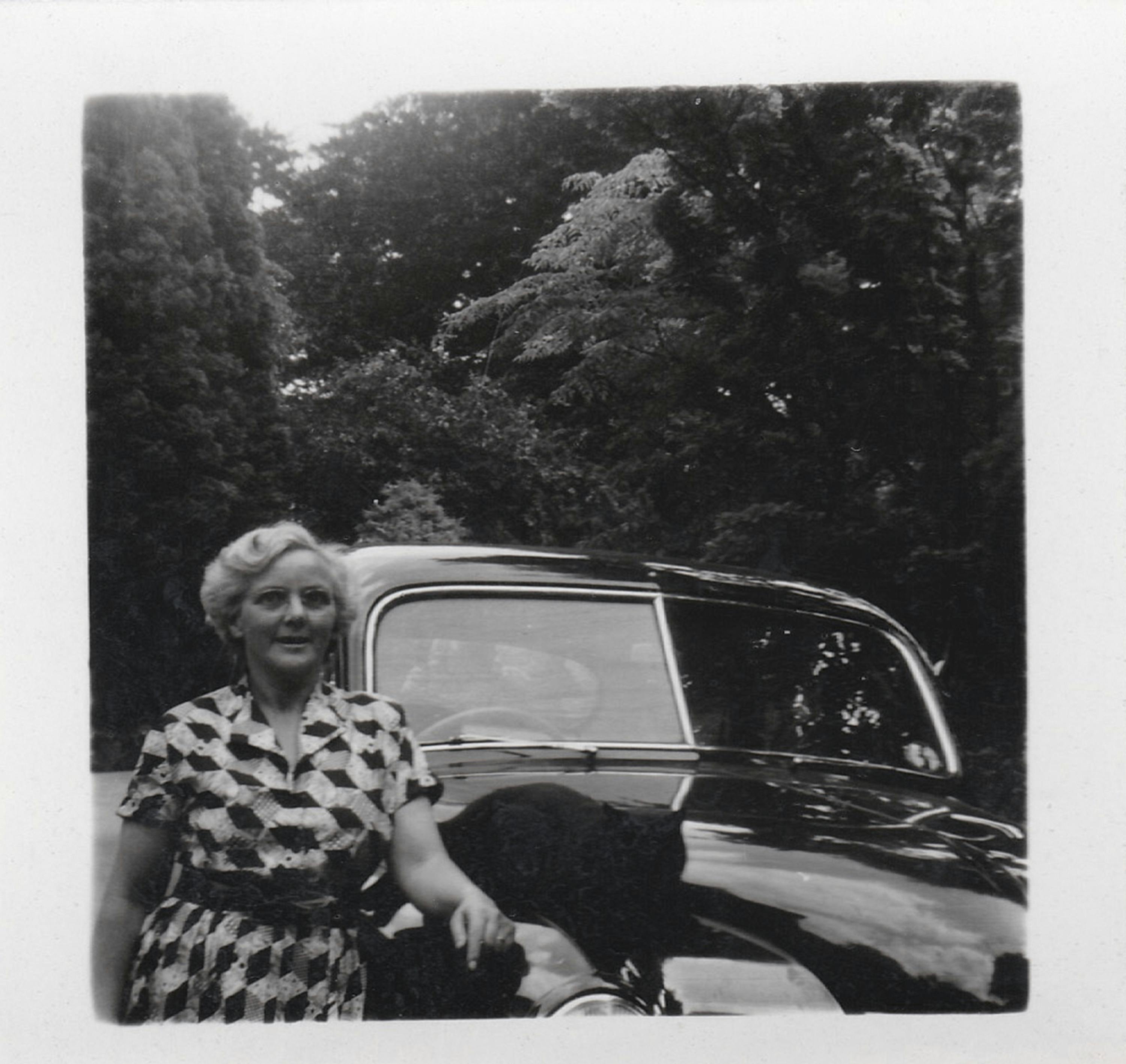 Black and white vintage photo of a woman standing by a classic car outdoors.