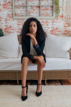 Fashionable woman in black blazer and heels sitting on a sofa in a modern interior.