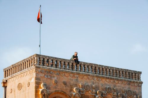 Free A woman sits atop a historic building featuring intricate stonework and a flag, under a clear sky. Stock Photo