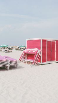 A vibrant pink beach hut and stacked sun loungers on a serene sandy beach, perfect for relaxation.