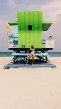 Woman in bikini celebrating in front of a colorful lifeguard tower on a sunny beach day.