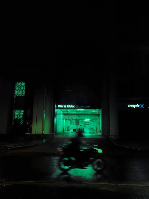 Free Silhouette of a motorcycle passing a neon-lit parking garage at night, captured in an urban setting. Stock Photo