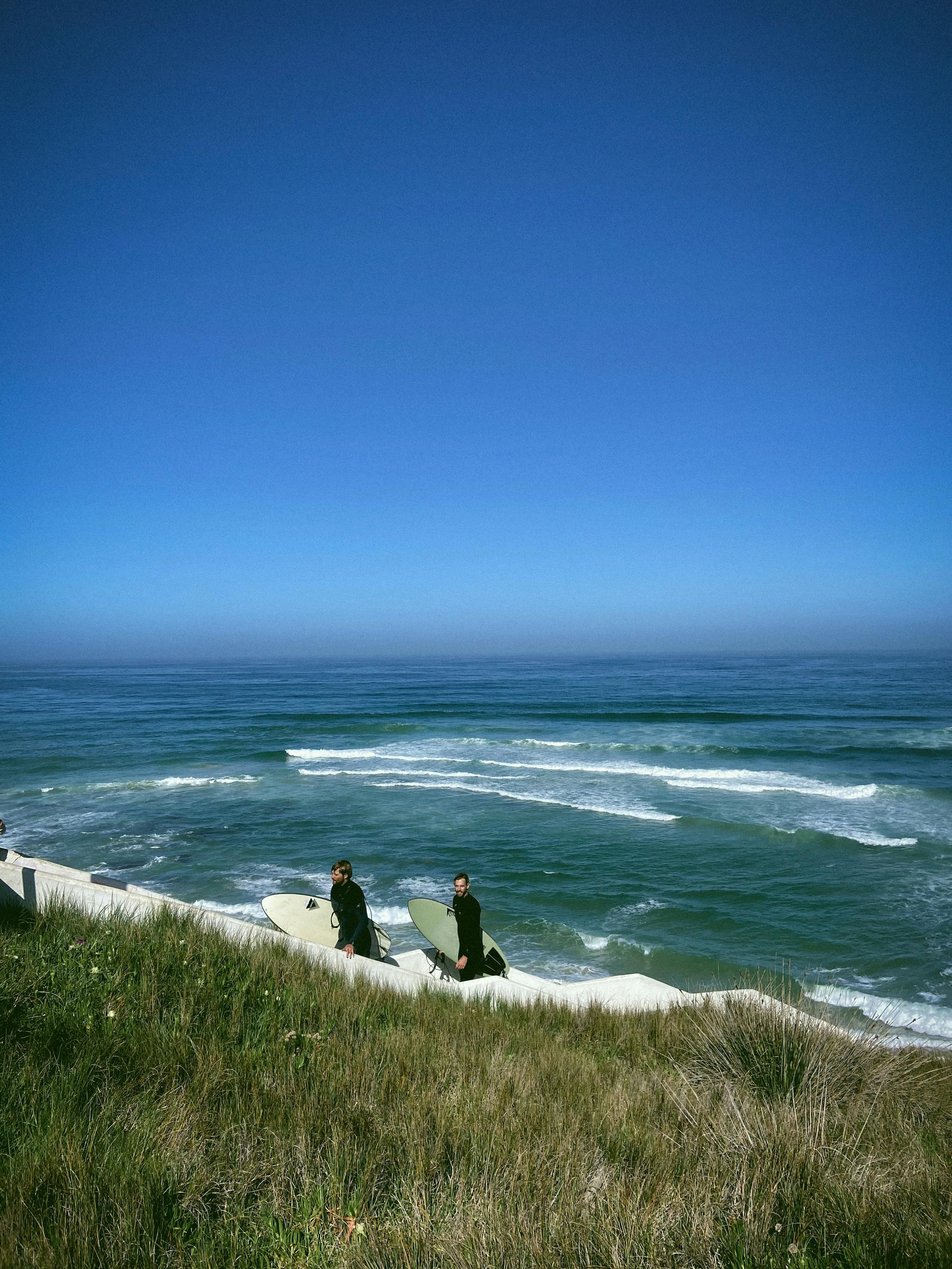 Two surfers in wetsuits walking along a sandy beach with surfboards, captured under a clear blue sky.