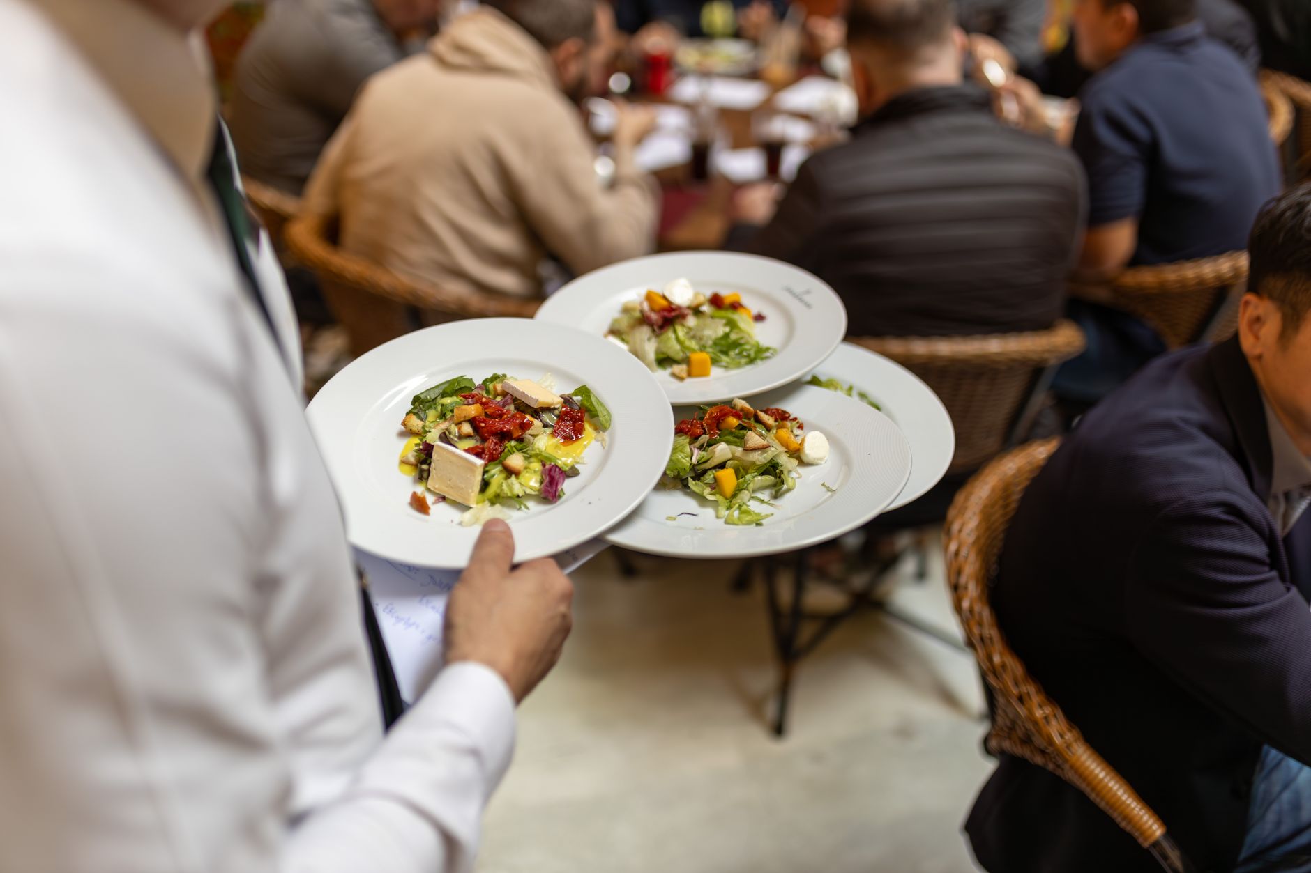 Waiter carrying plates with fresh salads in a busy restaurant setting.