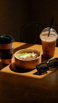 A warm breakfast scene with coffee, a smoothie, bowl of kiwi, granola, and sunglasses on a wooden table.