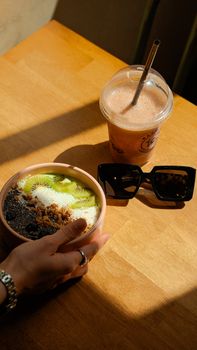 A vibrant breakfast bowl with fresh kiwi, paired with a smoothie and sunglasses on a wooden table.