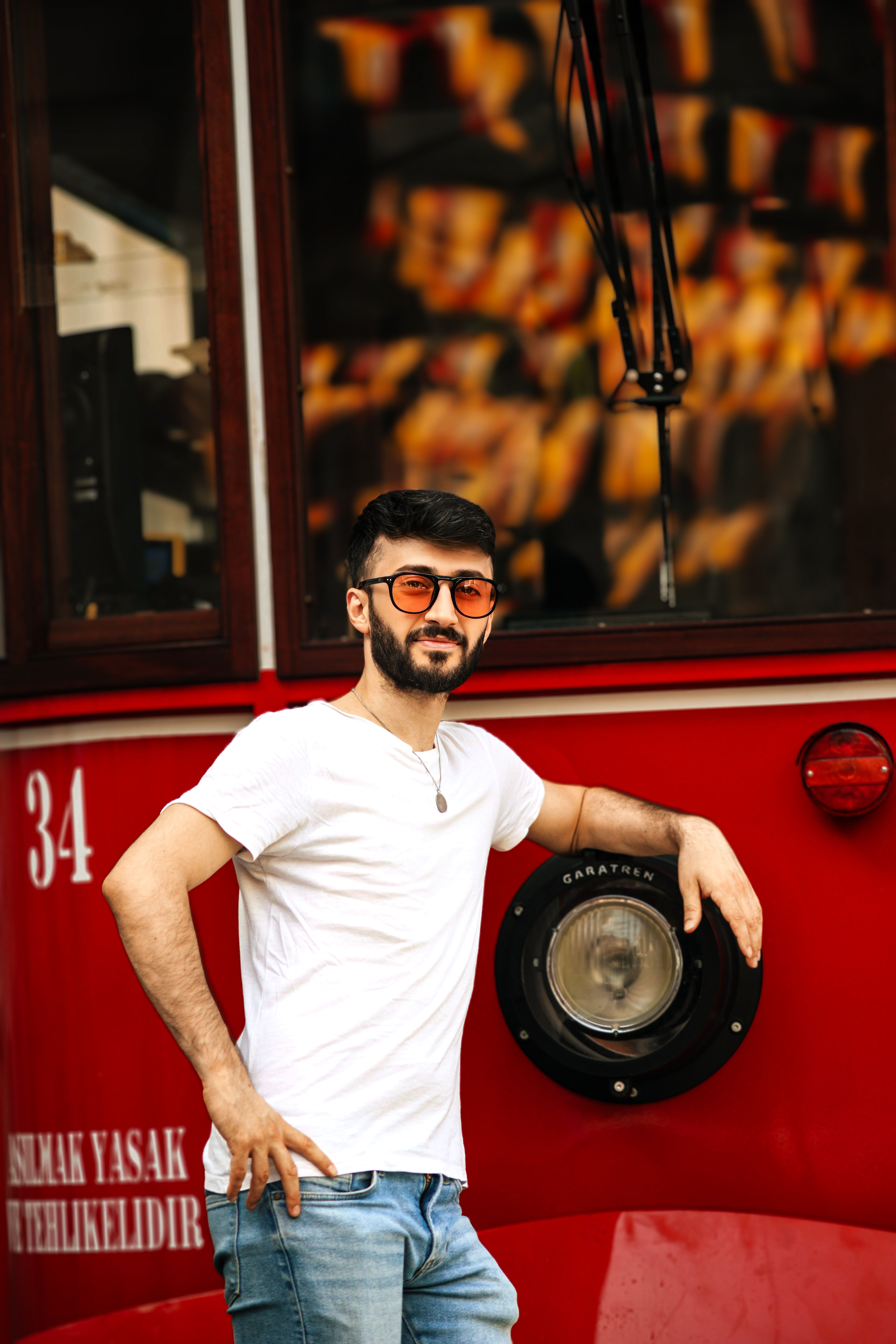 A fashionable man with eyeglasses poses by a classic red Istanbul tram.