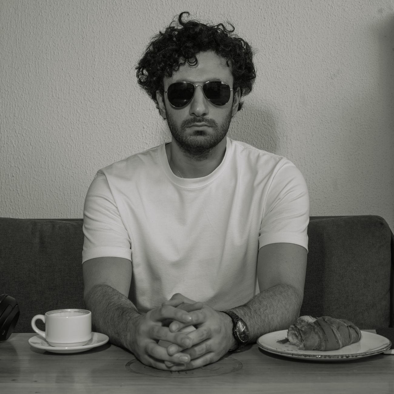 Stylish man in black and white portrait wearing sunglasses, seated with coffee and pastry.