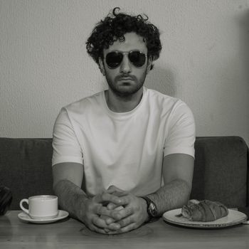 Stylish man in black and white portrait wearing sunglasses, seated with coffee and pastry.