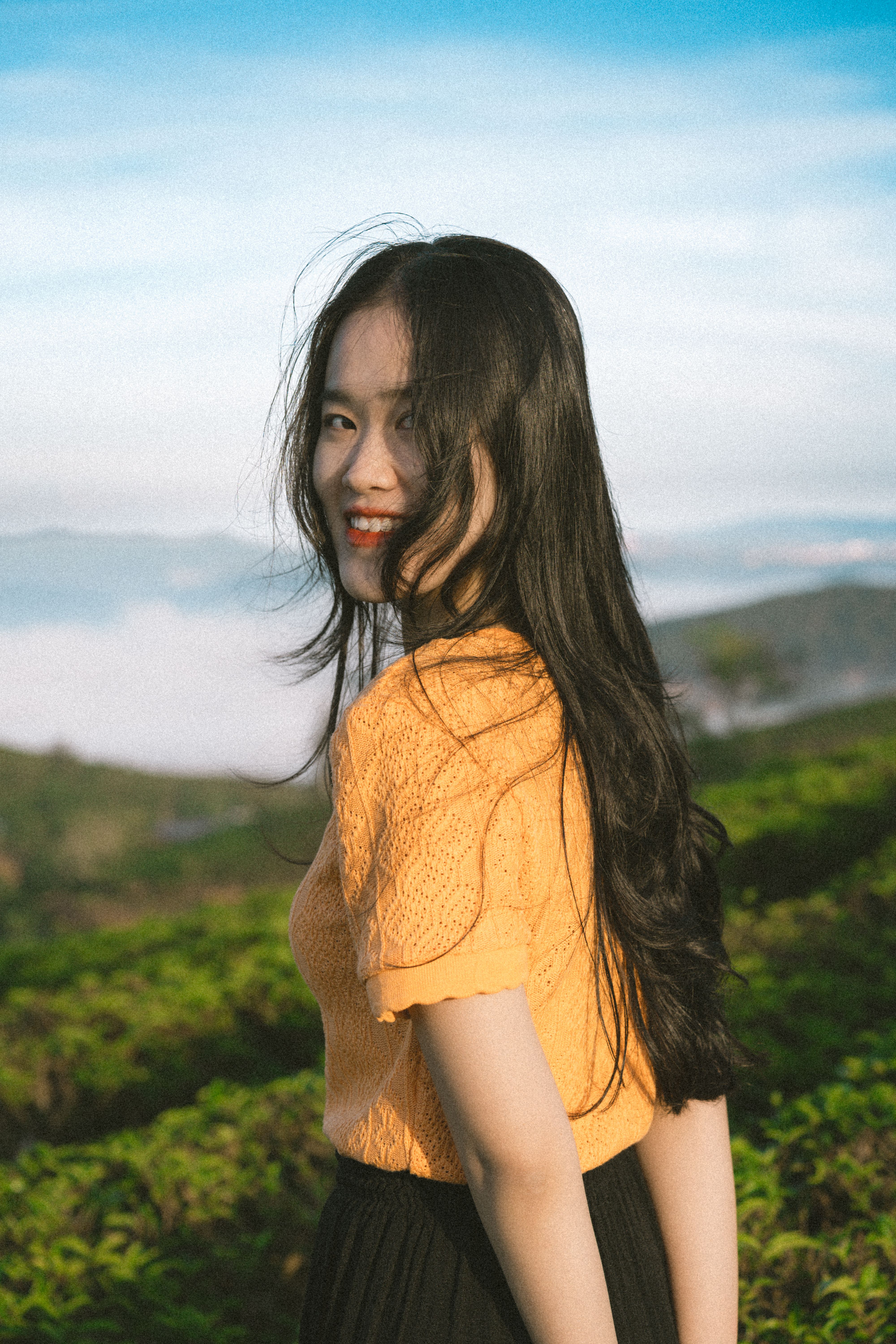 Asian woman in orange blouse smiling against scenic Dalat mountains.