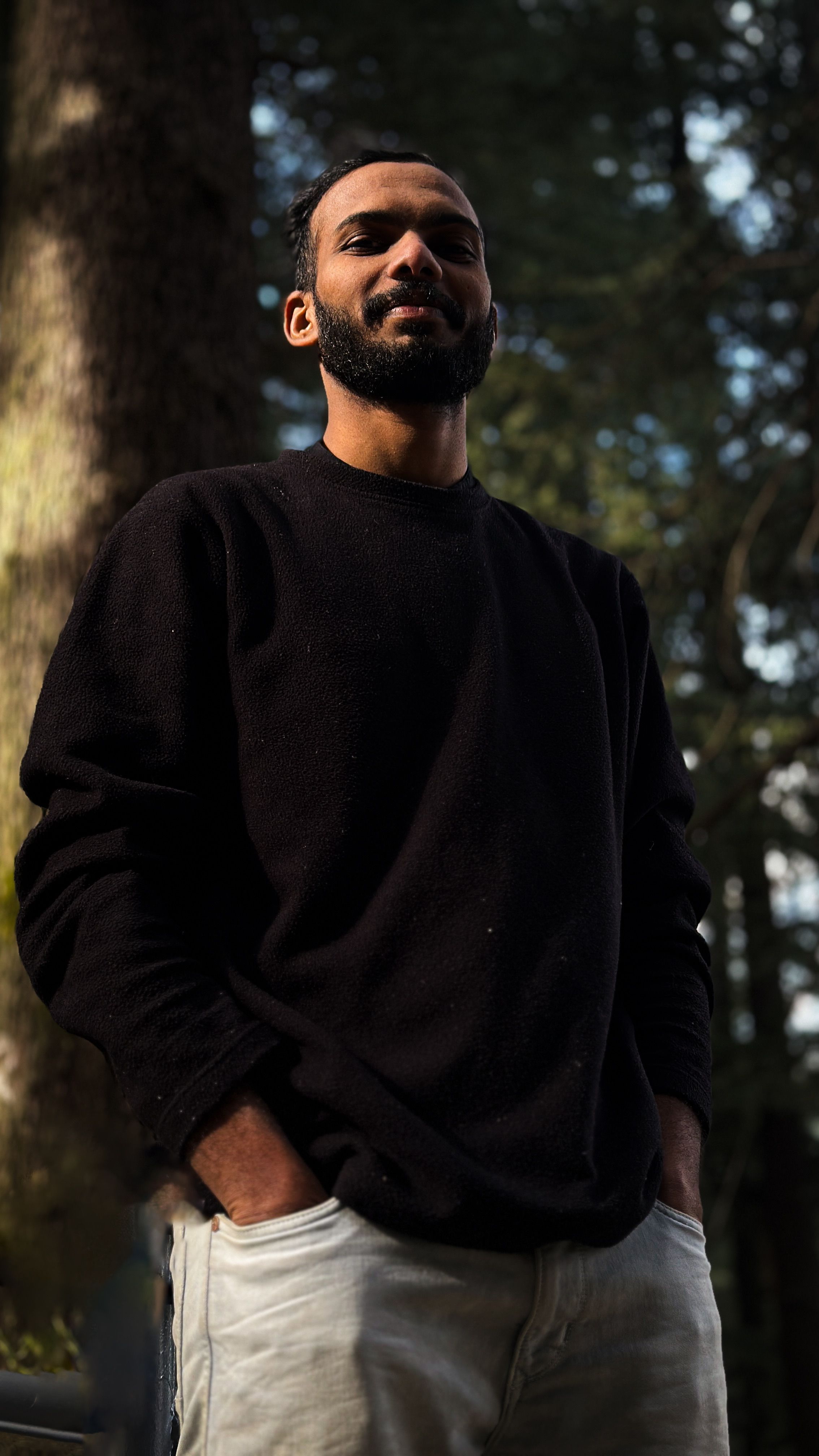 Portrait of a smiling man standing outdoors with hands in pockets, embracing a serene nature setting.