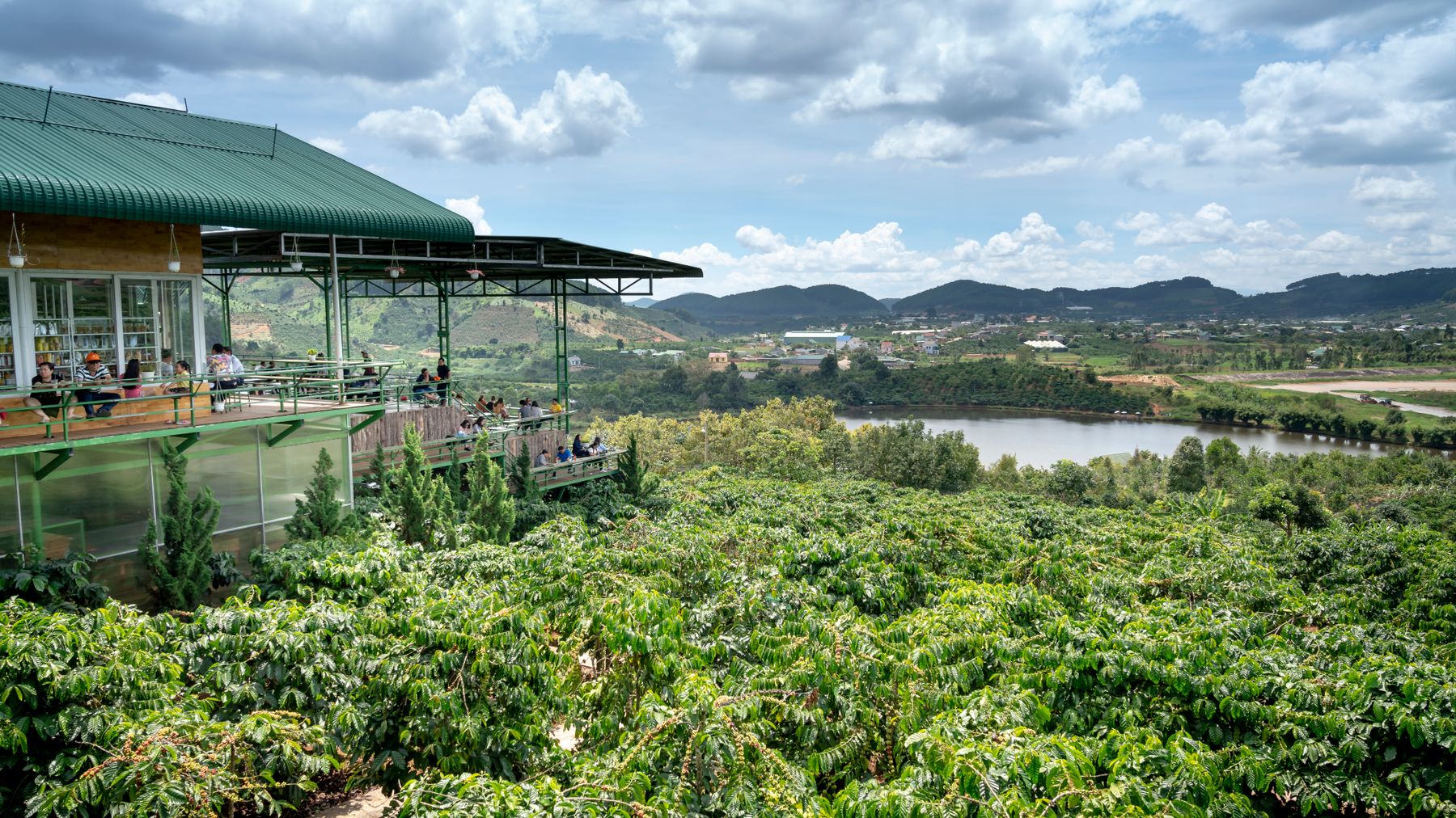 A picturesque cafe situated above lush tropical farms and a serene pond, framed by hilly landscapes under a cloudy sky.