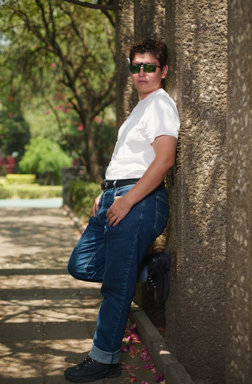 A man leaning against a tree wearing jeans and a white shirt