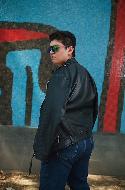 A man in a leather jacket and sunglasses standing in front of a wall