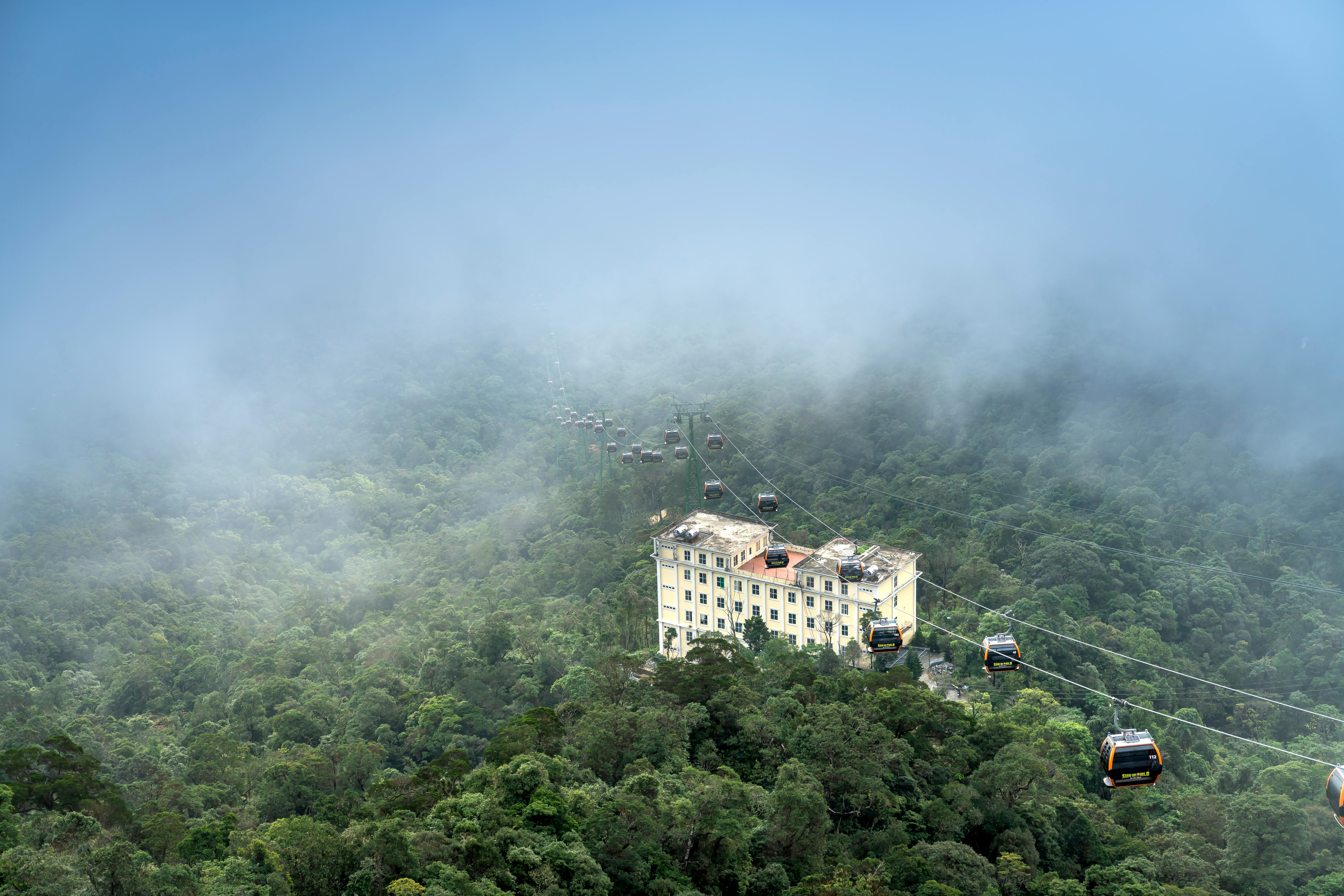 缆车行驶在云雾缭绕的山间森林上空，鸟瞰图中可见一栋建筑