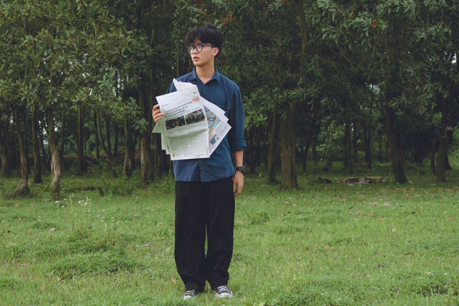 A young man stands in a green park holding newspapers, surrounded by trees and grass.