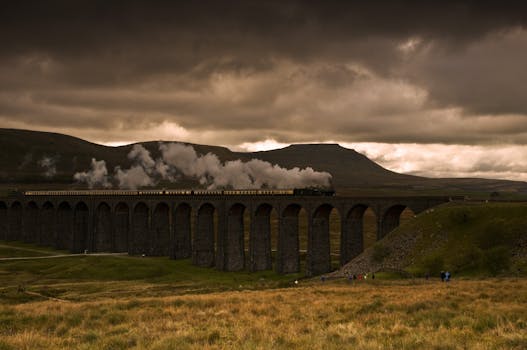 A steam train crosses a picturesque viaduct against a dramatic sky.