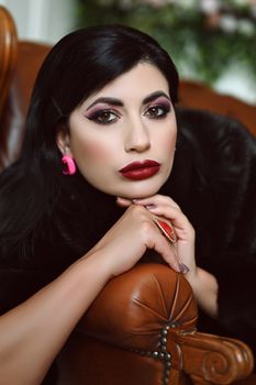 Portrait of a woman with red lipstick and stylish makeup, leaning on a leather chair indoors.