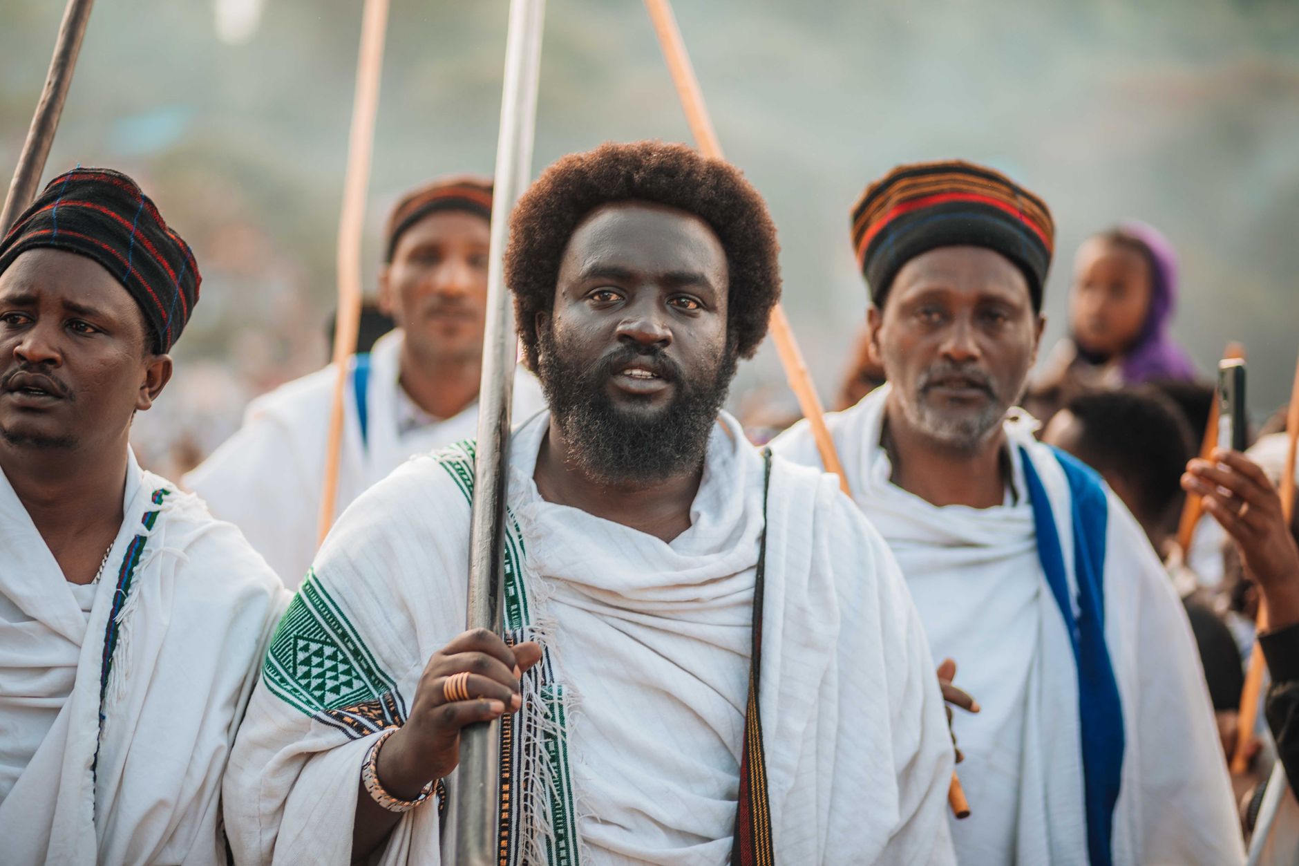 Men wearing traditional robes participate in a cultural event, holding wooden staffs.