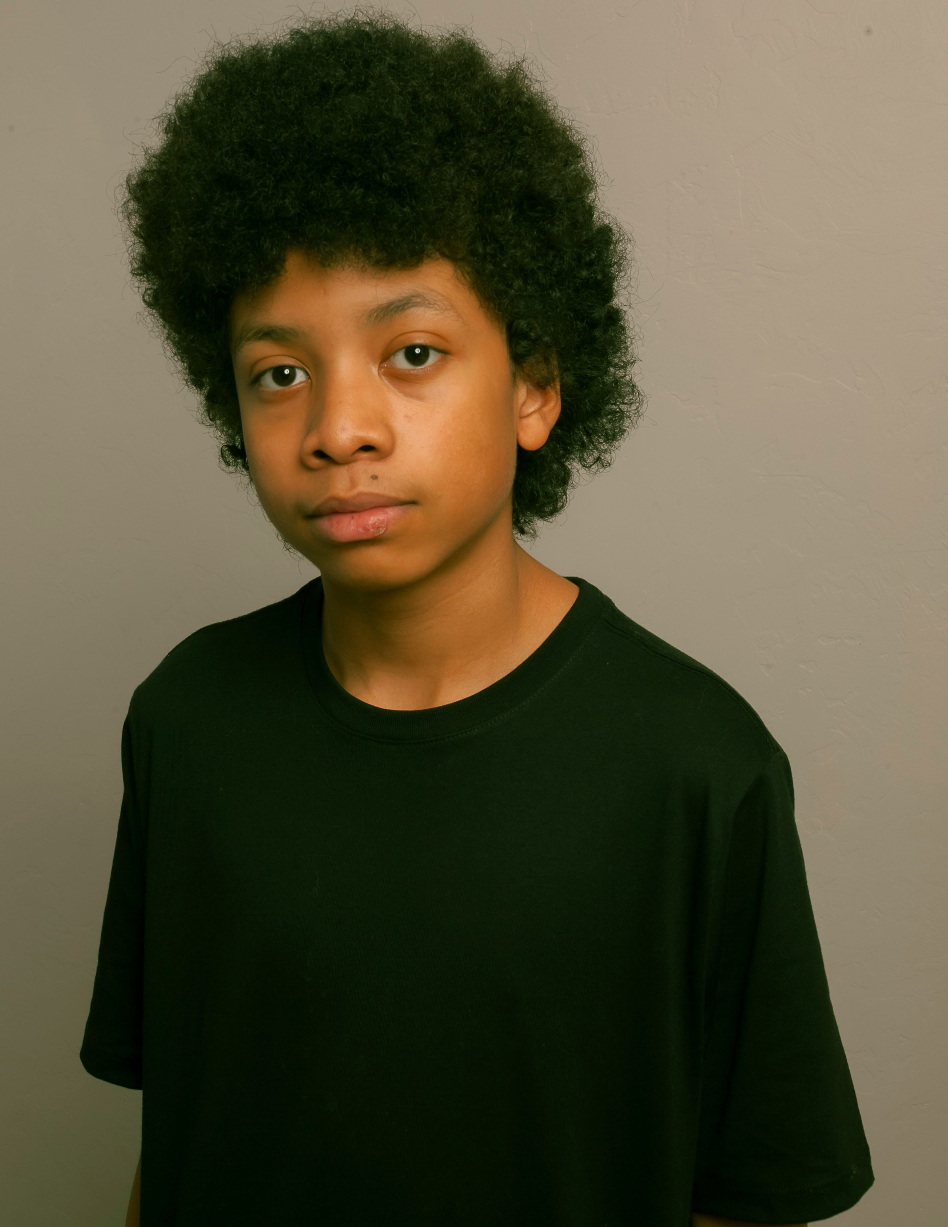 Studio portrait of a young boy with curly black hair wearing a black T-shirt against a gray background.