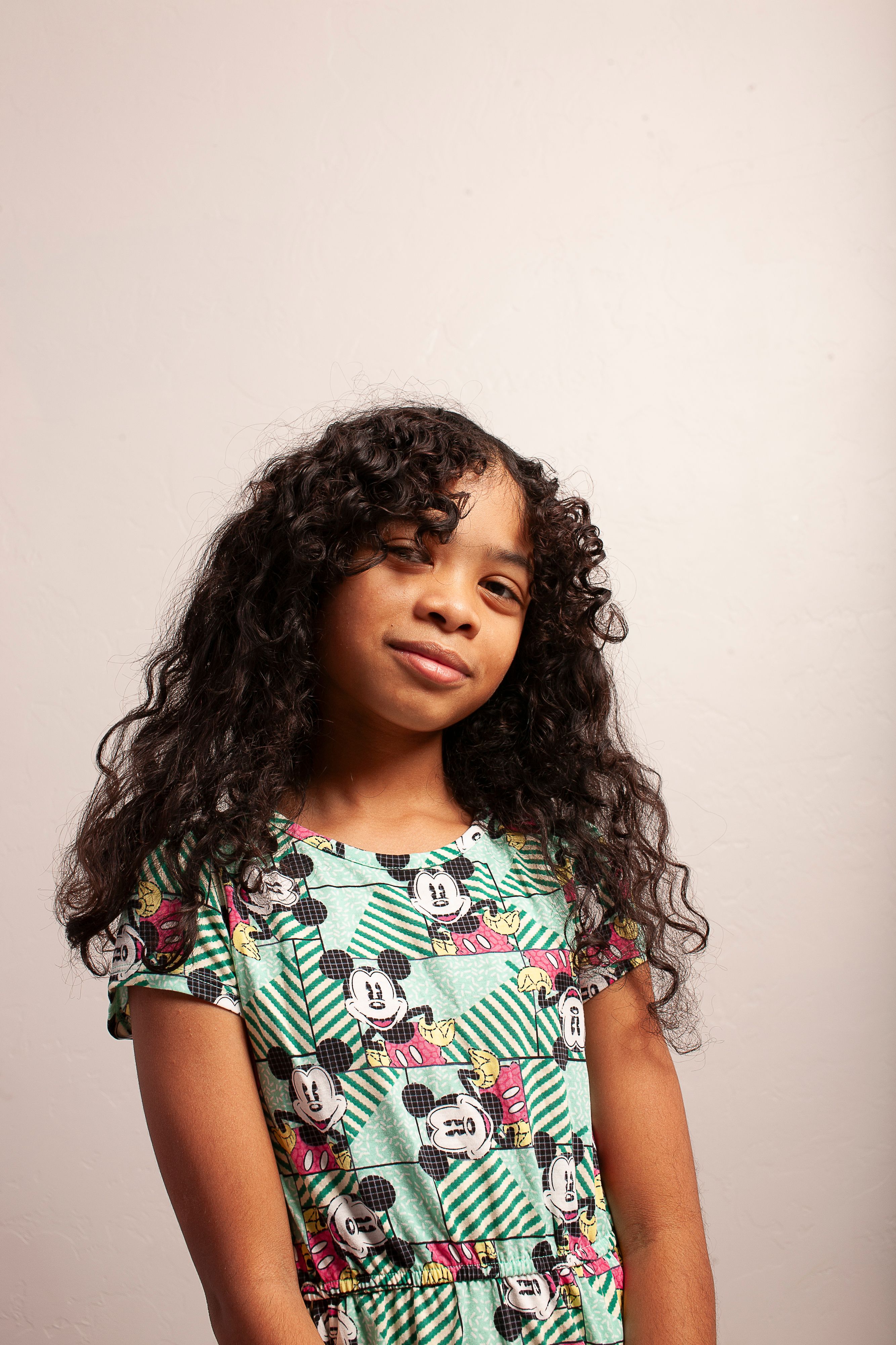 Studio portrait of a young girl with long curly hair wearing a colorful dress, smiling softly.