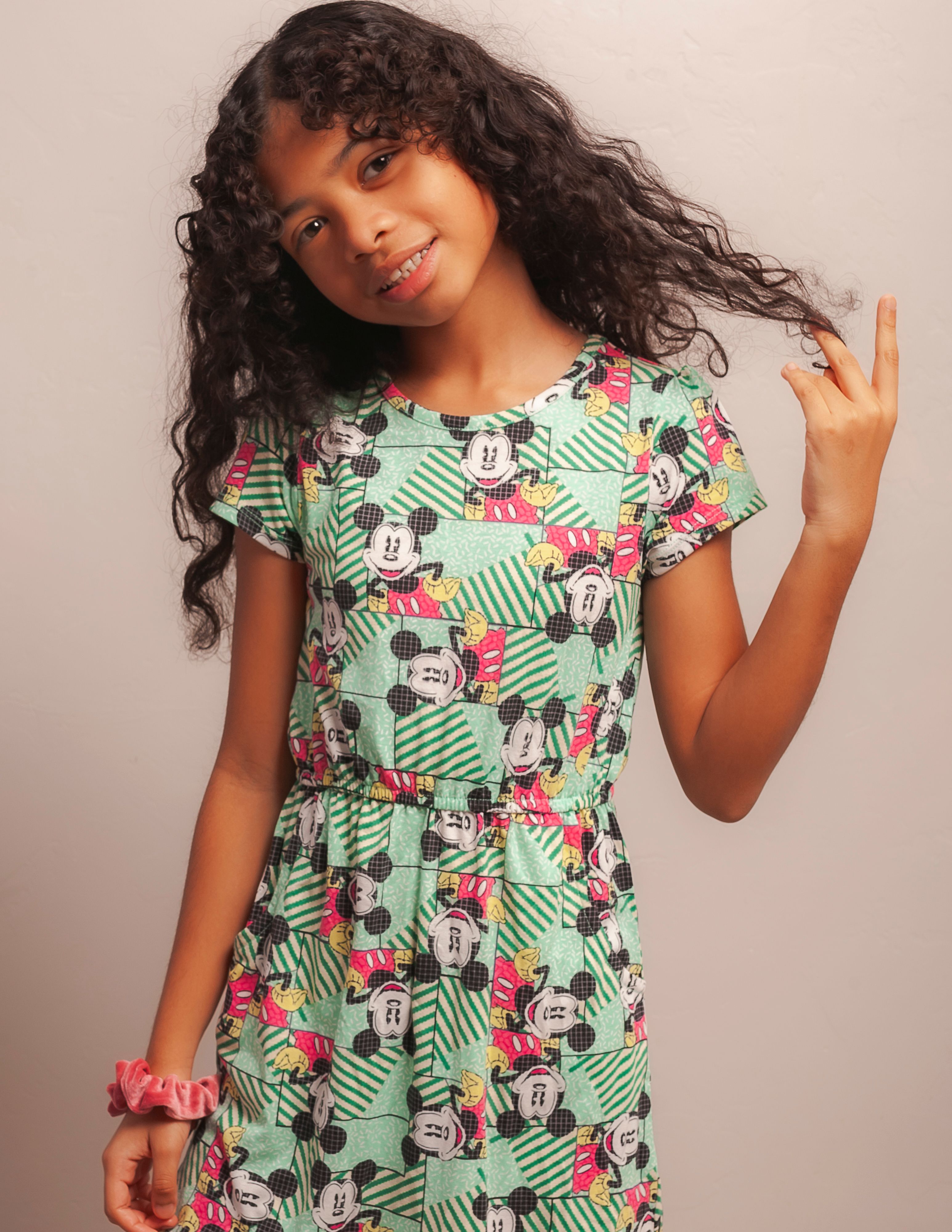 Portrait of a cheerful teenage girl in a bright Mickey Mouse sundress, posing indoors with a playful touch to her hair.