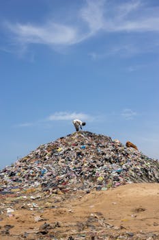 A man stands atop a mound of garbage under a bright blue sky, highlighting pollution.