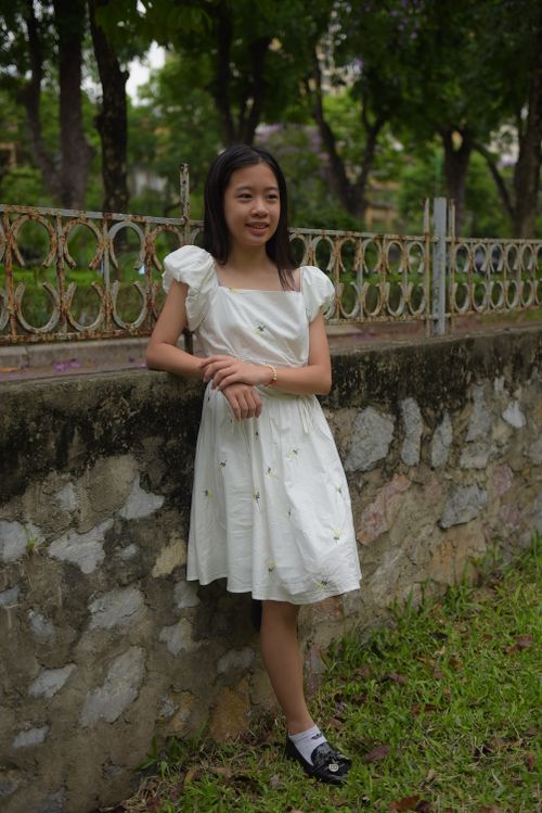 Free A young Asian teen girl in a white dress leans against a stone wall in a park setting. Stock Photo