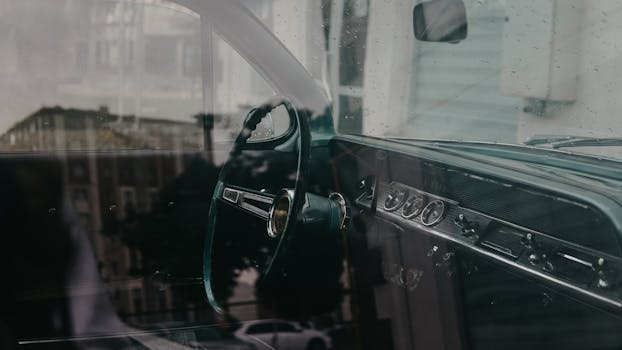 A close-up of a vintage car's dashboard and steering wheel with reflections and raindrops on the window.