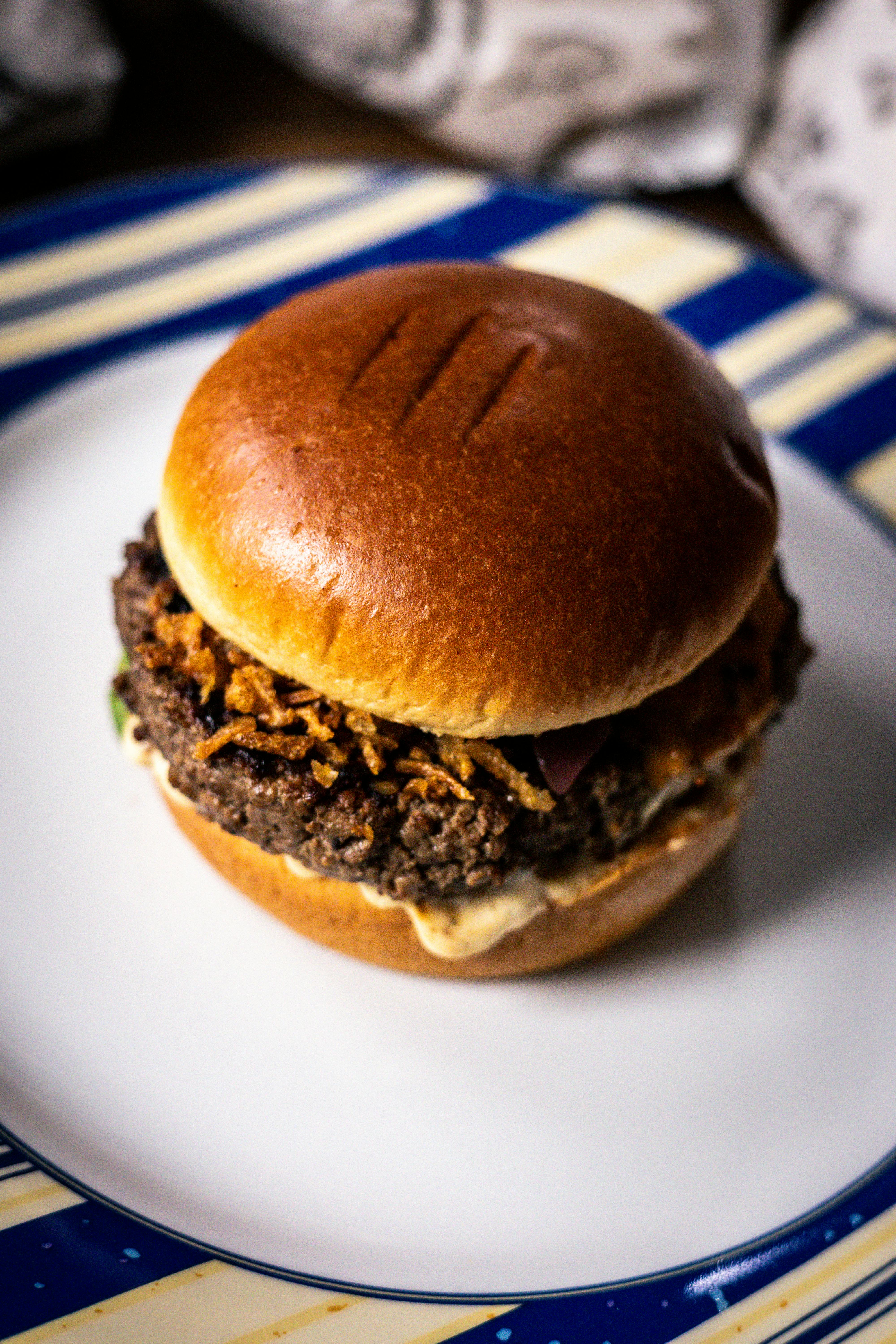 A glossy beef burger on a golden brioche bun, topped with white coconut sambal, golden fried shallots, and fresh green herbs, with dark, glossy rendang glaze coating the patty