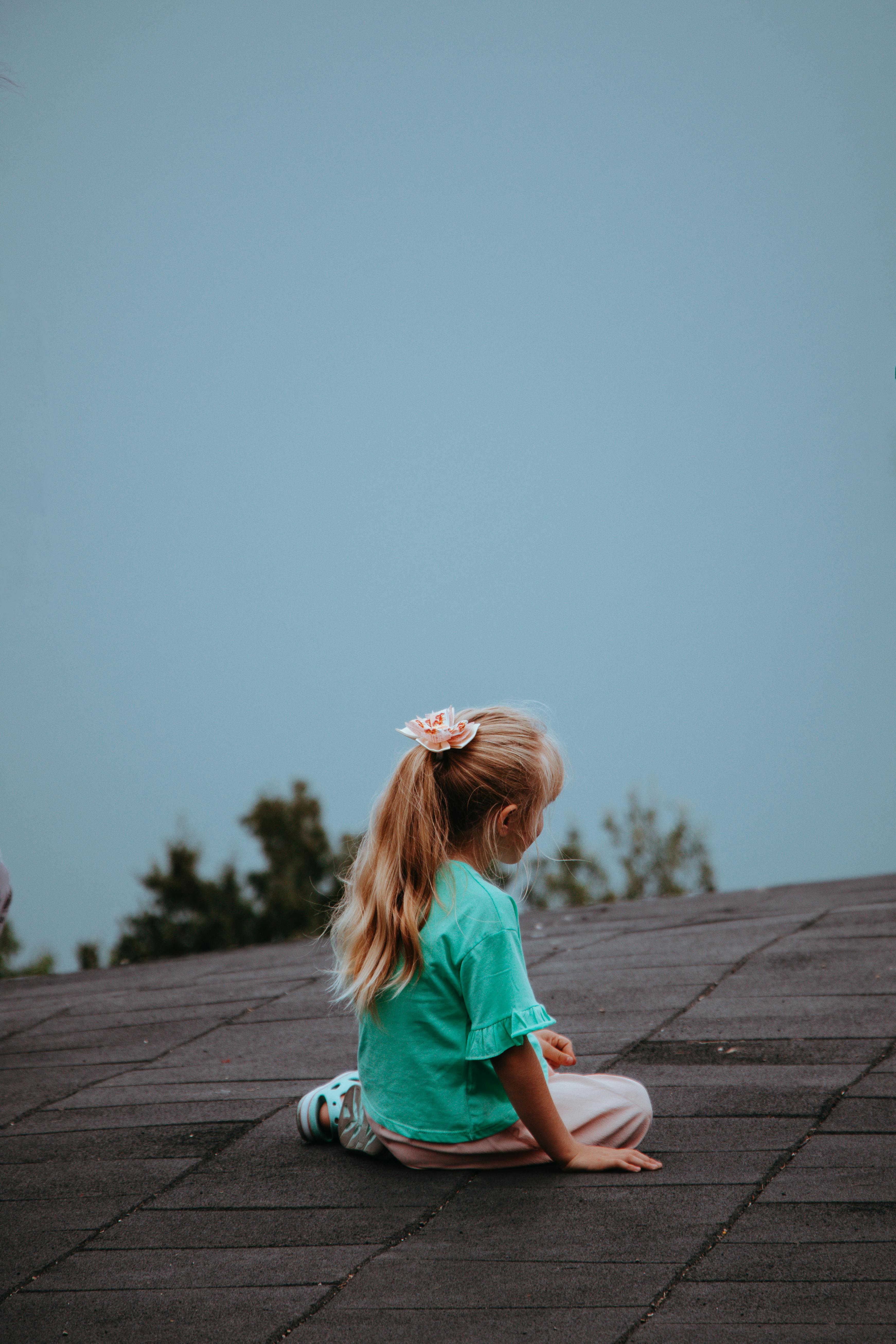 A blonde child sits peacefully on a rooftop with a clear sky backdrop, symbolizing innocence and serenity.