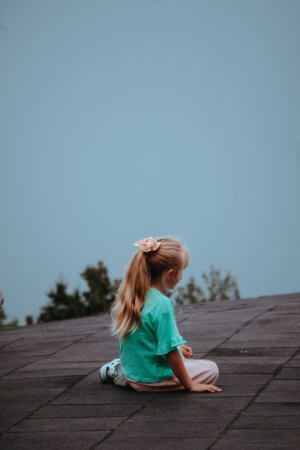 A blonde child sits peacefully on a rooftop with a clear sky backdrop, symbolizing innocence and serenity.