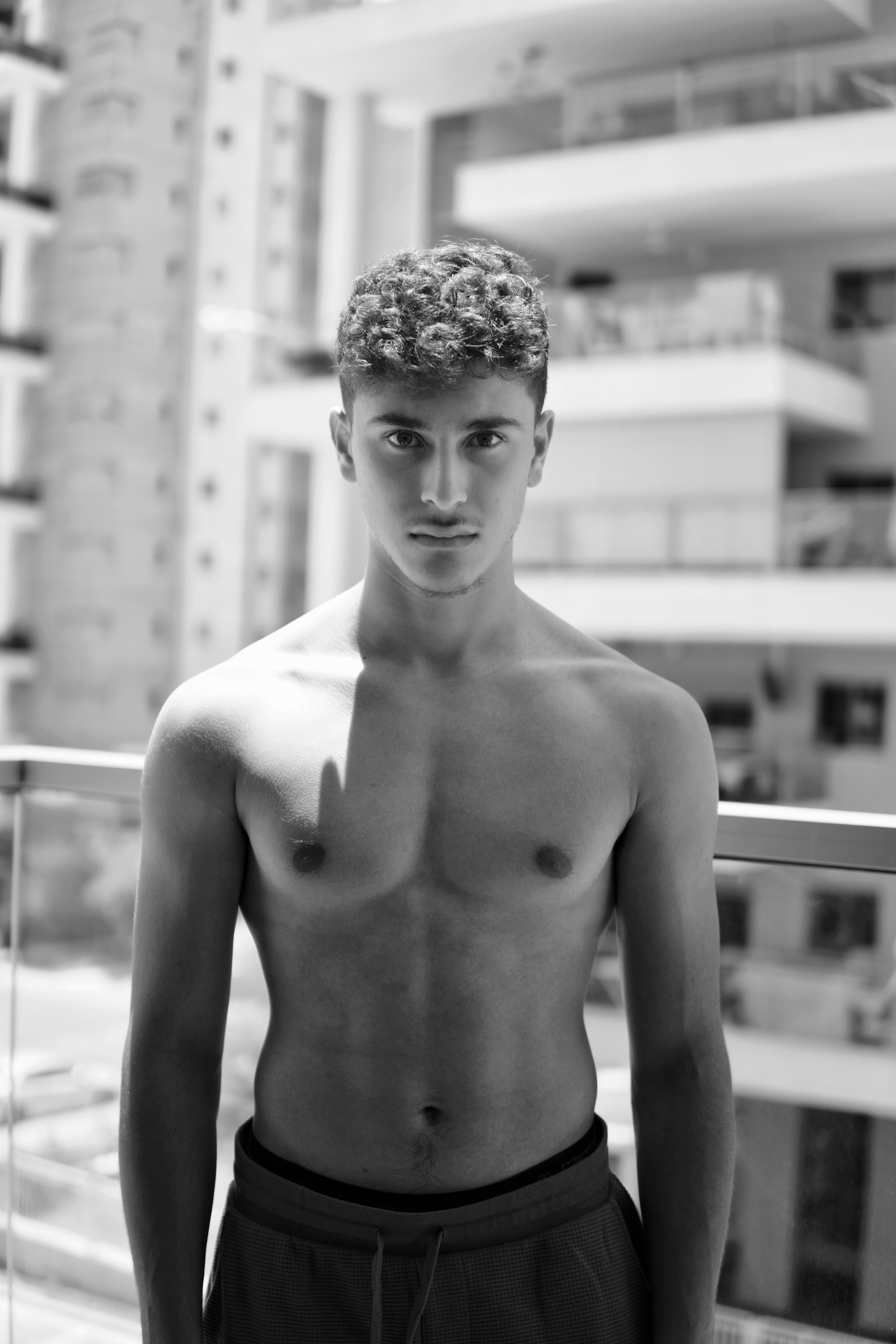Striking black and white portrait of a shirtless man standing on a balcony with urban architecture background.