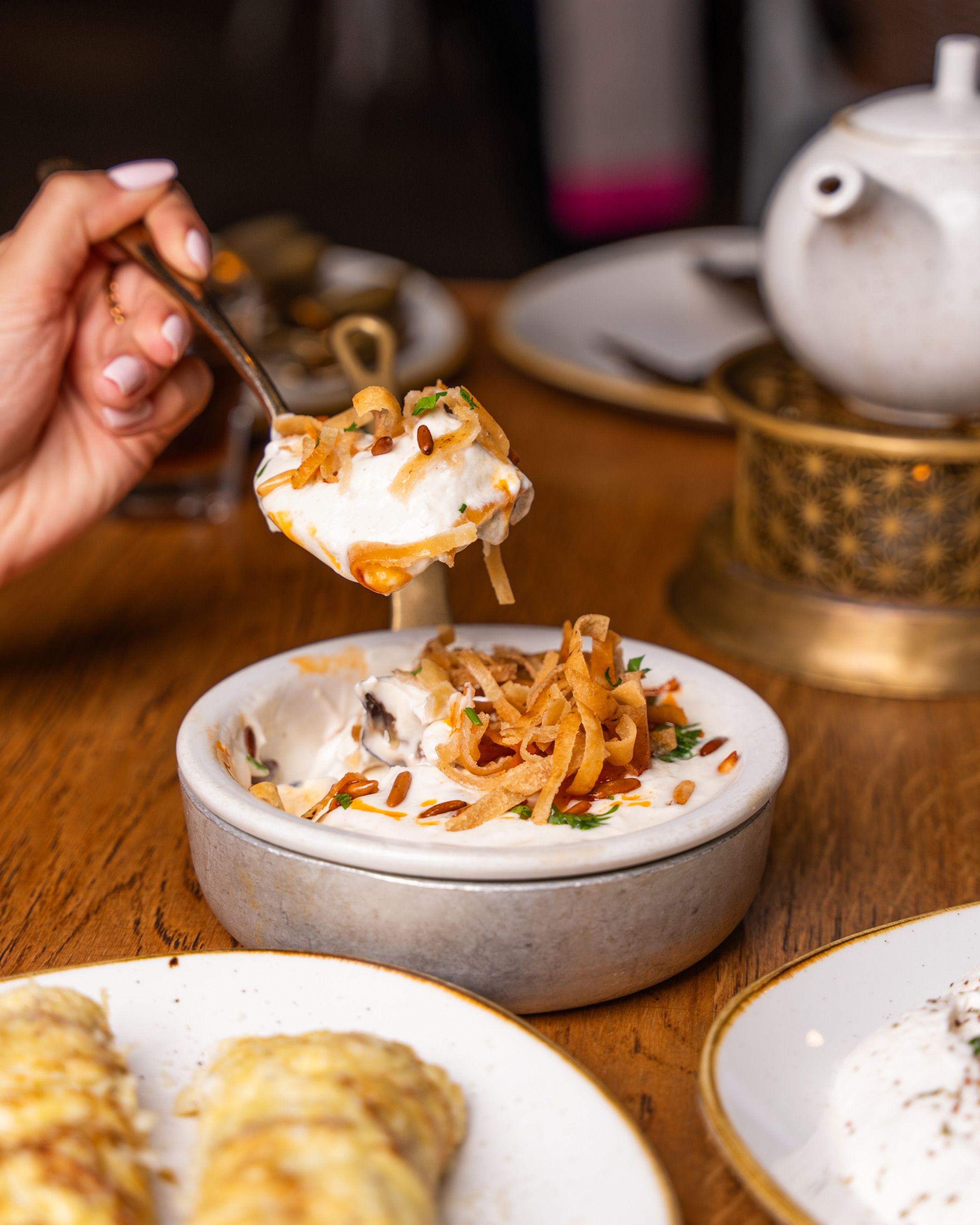 Close-up of a hand serving a creamy dish topped with fried onions and herbs in a cozy dining atmosphere.
