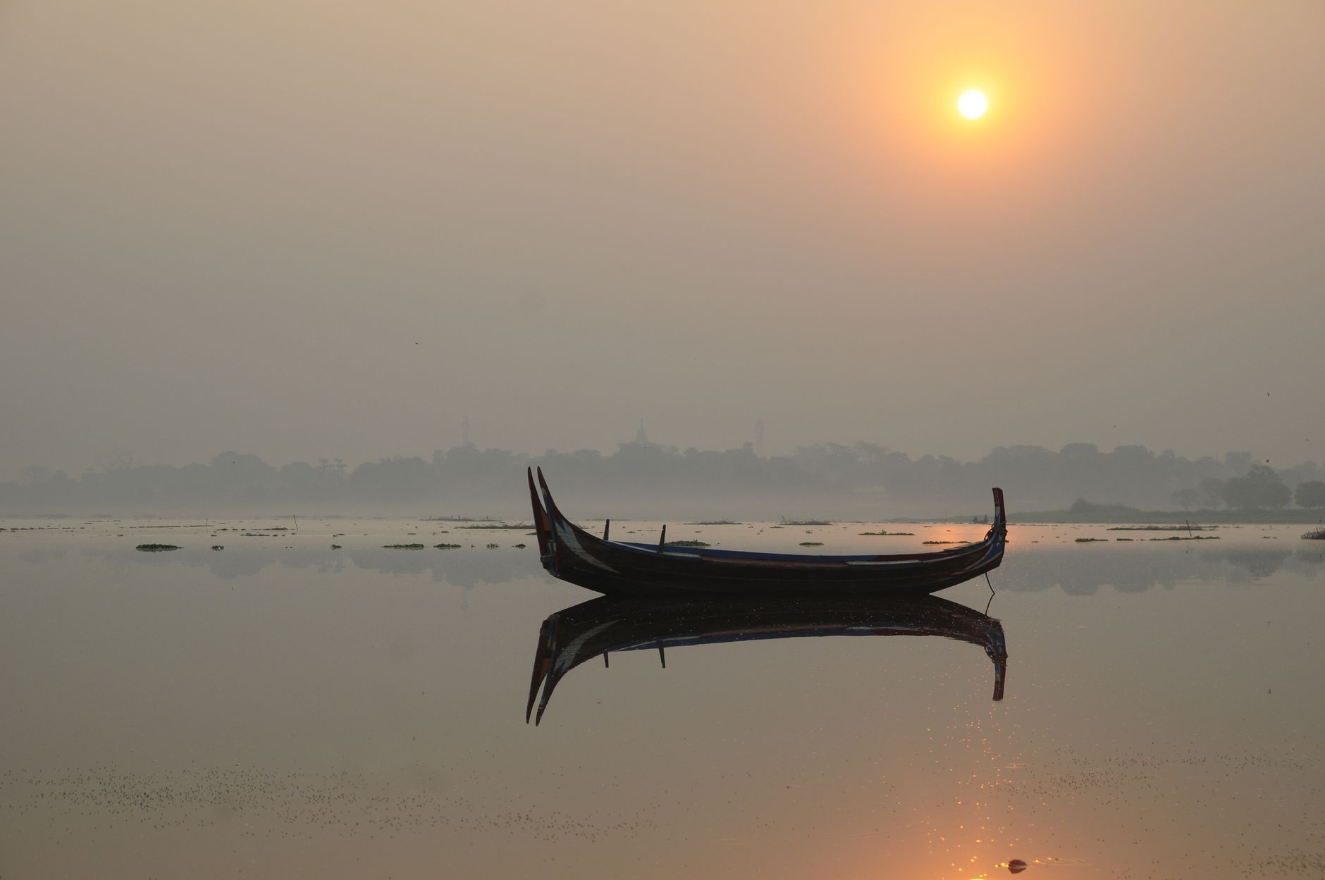 Peaceful sunrise reflecting over a calm lake with a silhouetted canoe in Mandalay, Myanmar.