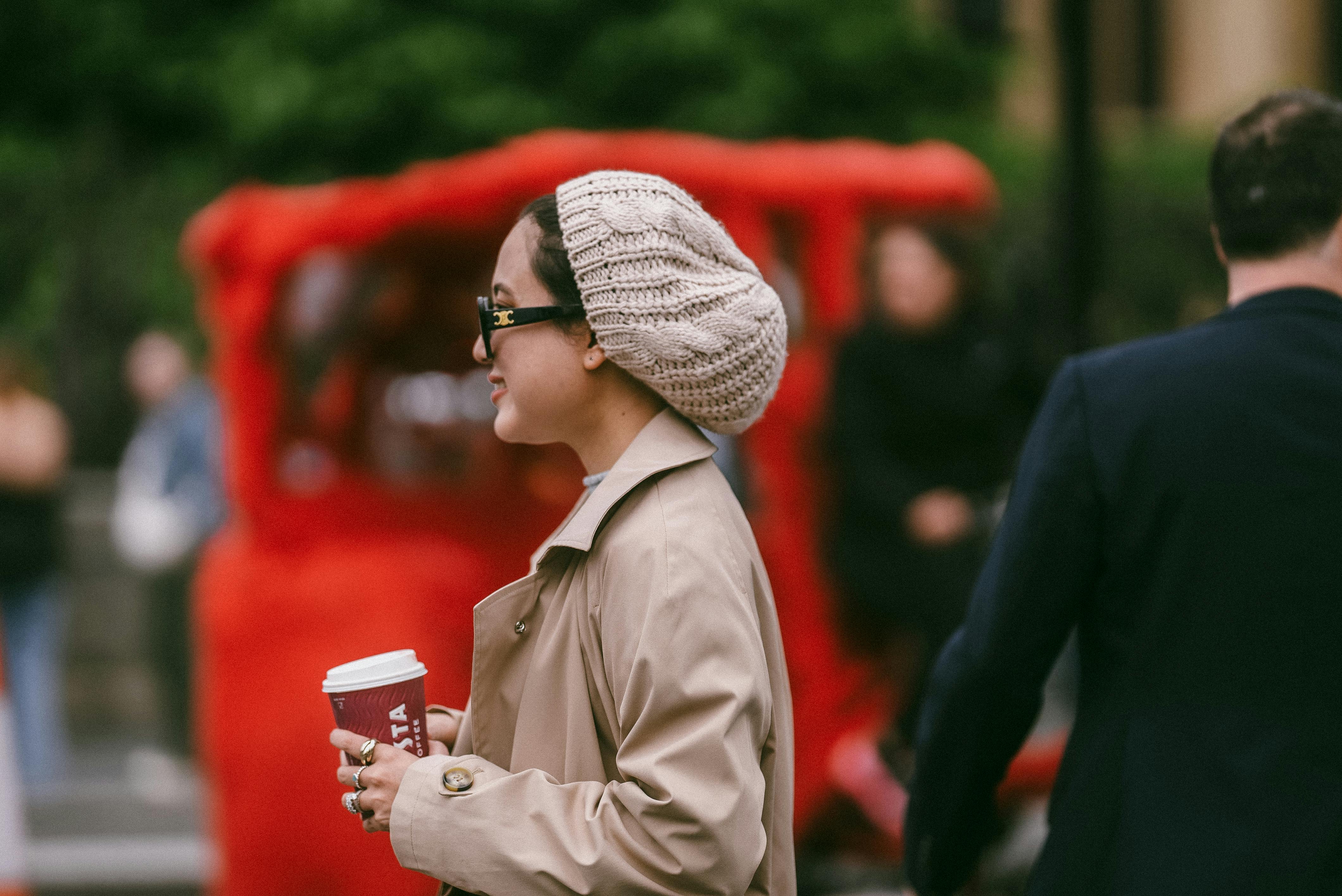 A stylish woman with a coffee cup in a bustling city street, wearing a knit hat and trench coat.
