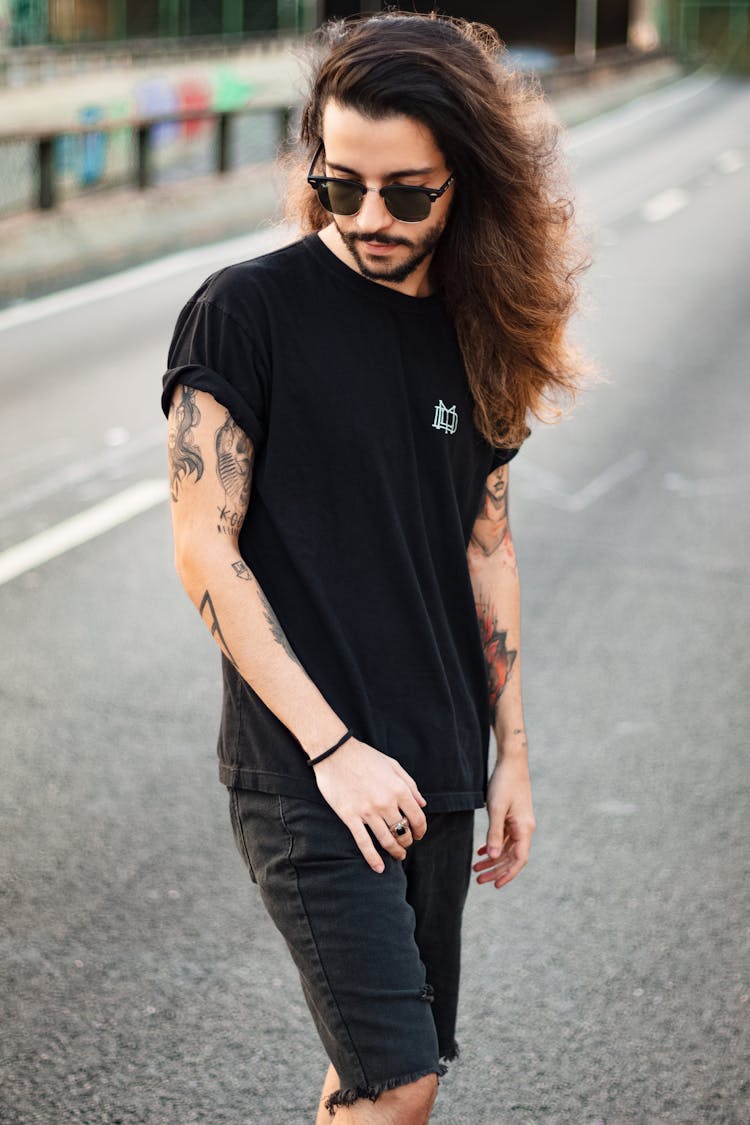 Photo Of Tattooed Man In Black T-shirt, Sunglasses, And Black Denim Shorts Posing In Middle Of Road While Looking Down