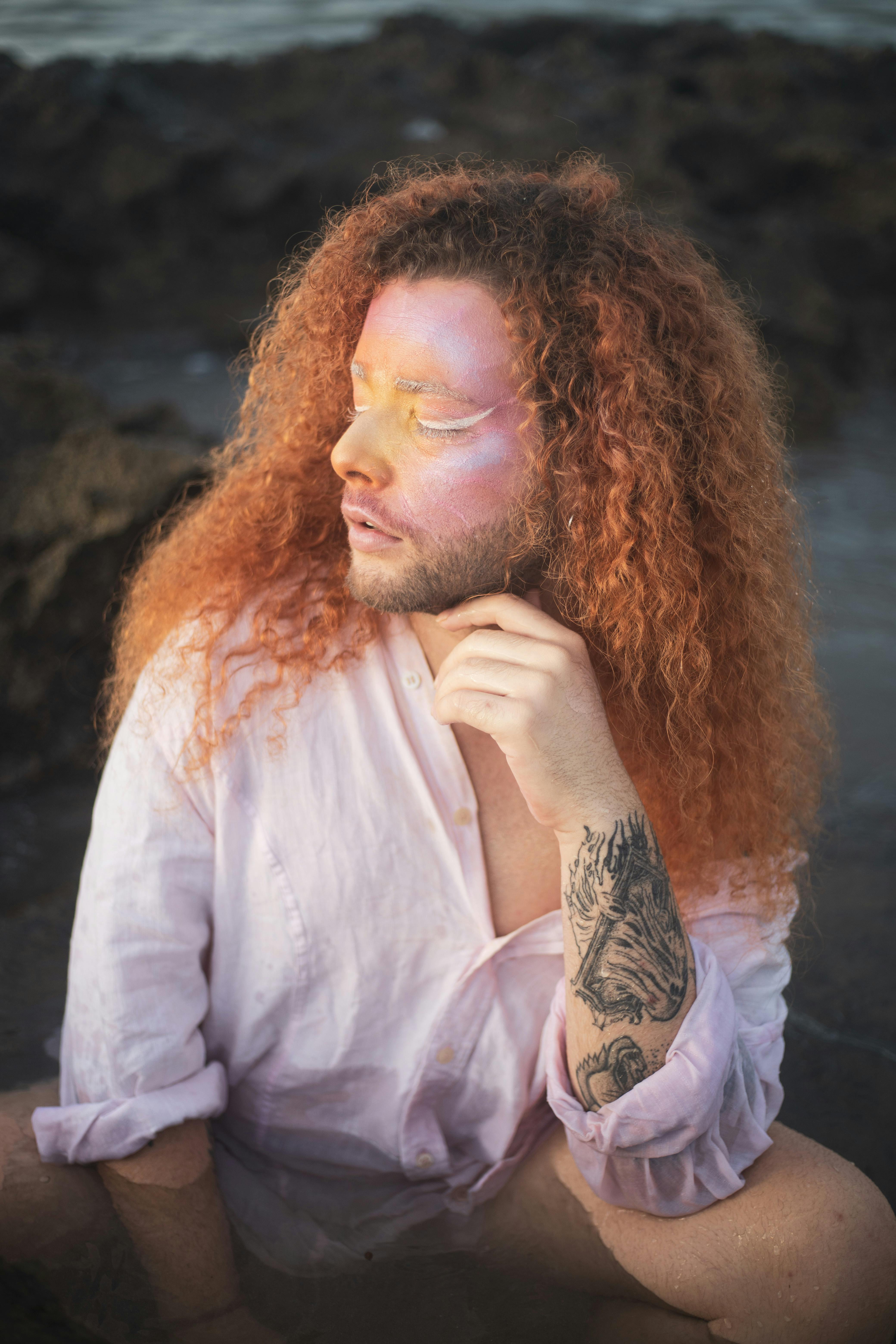 Portrait of a red-haired man with artistic makeup and tattoos, posing outdoors by the water.