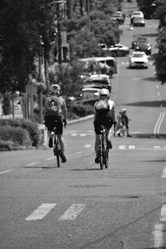 Black and white photo of cyclists riding bicycles through busy city streets, with cars and pedestrians.